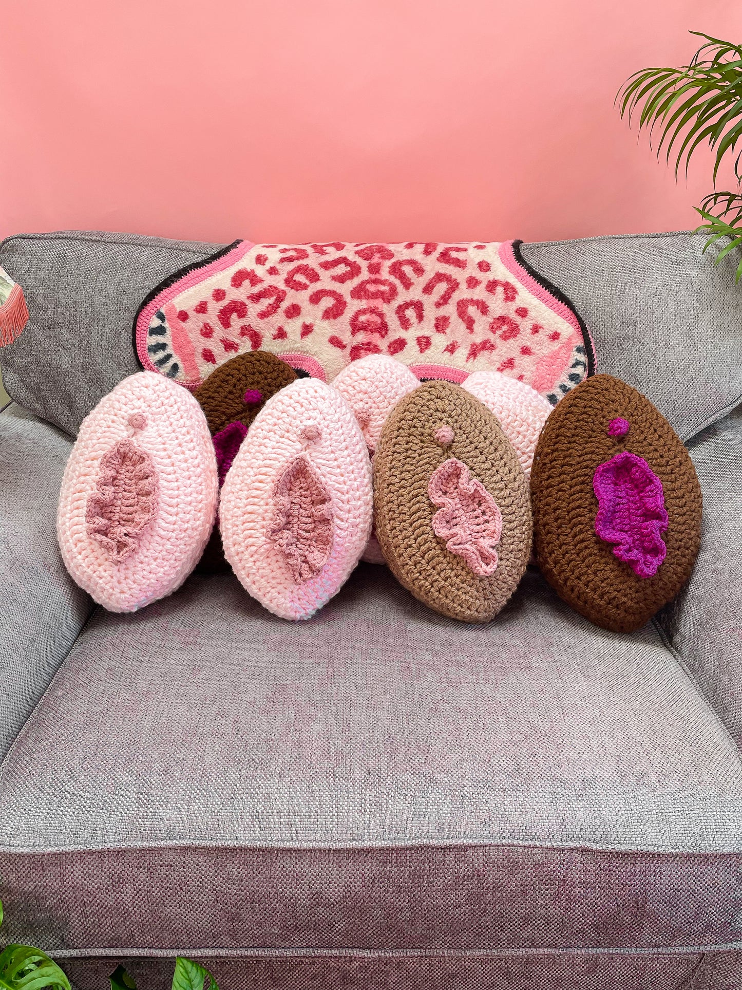 Seven vulva inspired pillows in various cream and brown colours lying together on a grey sofa with a pink leopard blanket behind them and plants around.