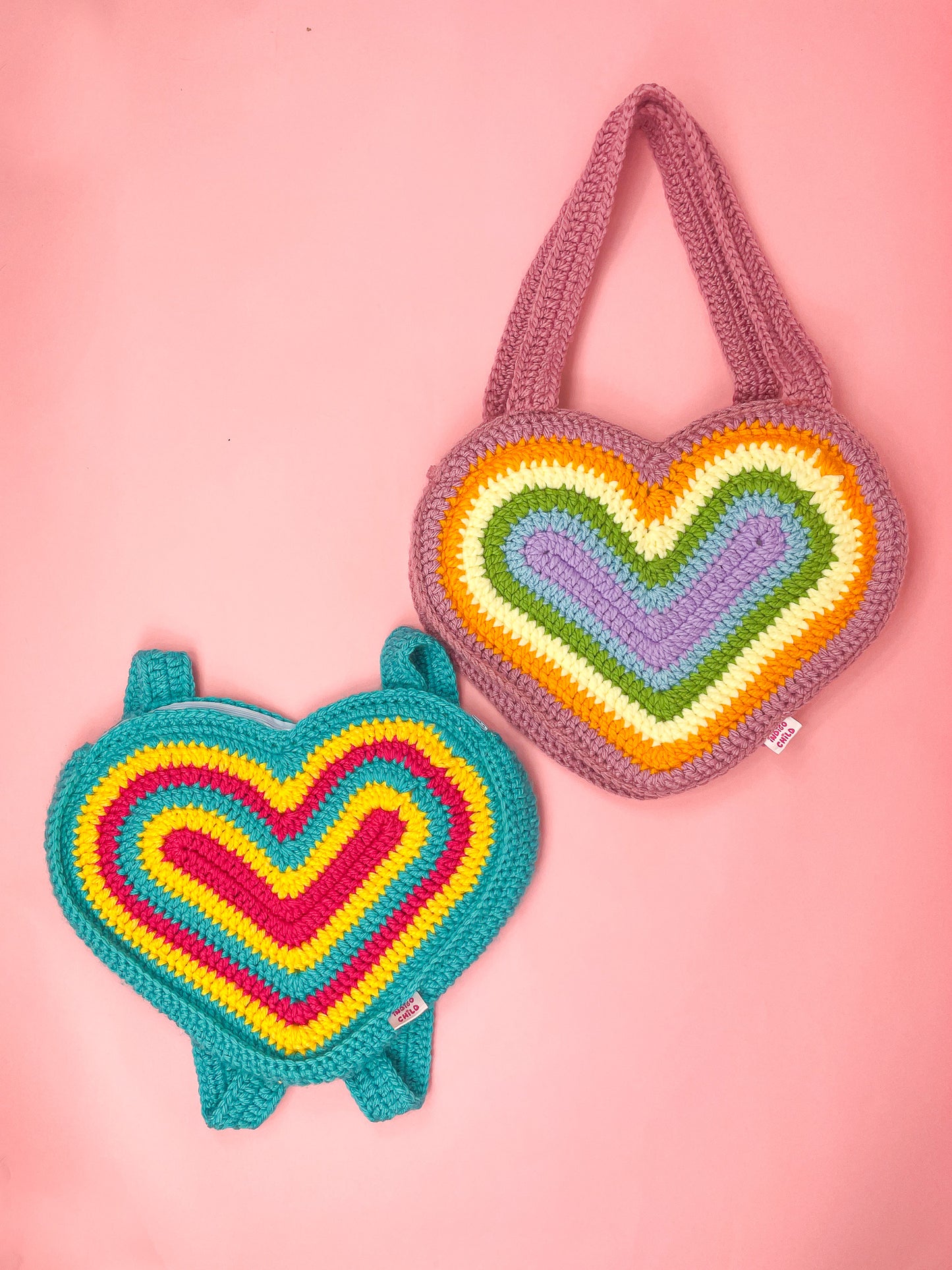 Two heart shaped bags lying together on a pink background, one in pansexual pride flag colours and the other in pastel rainbow colours.
