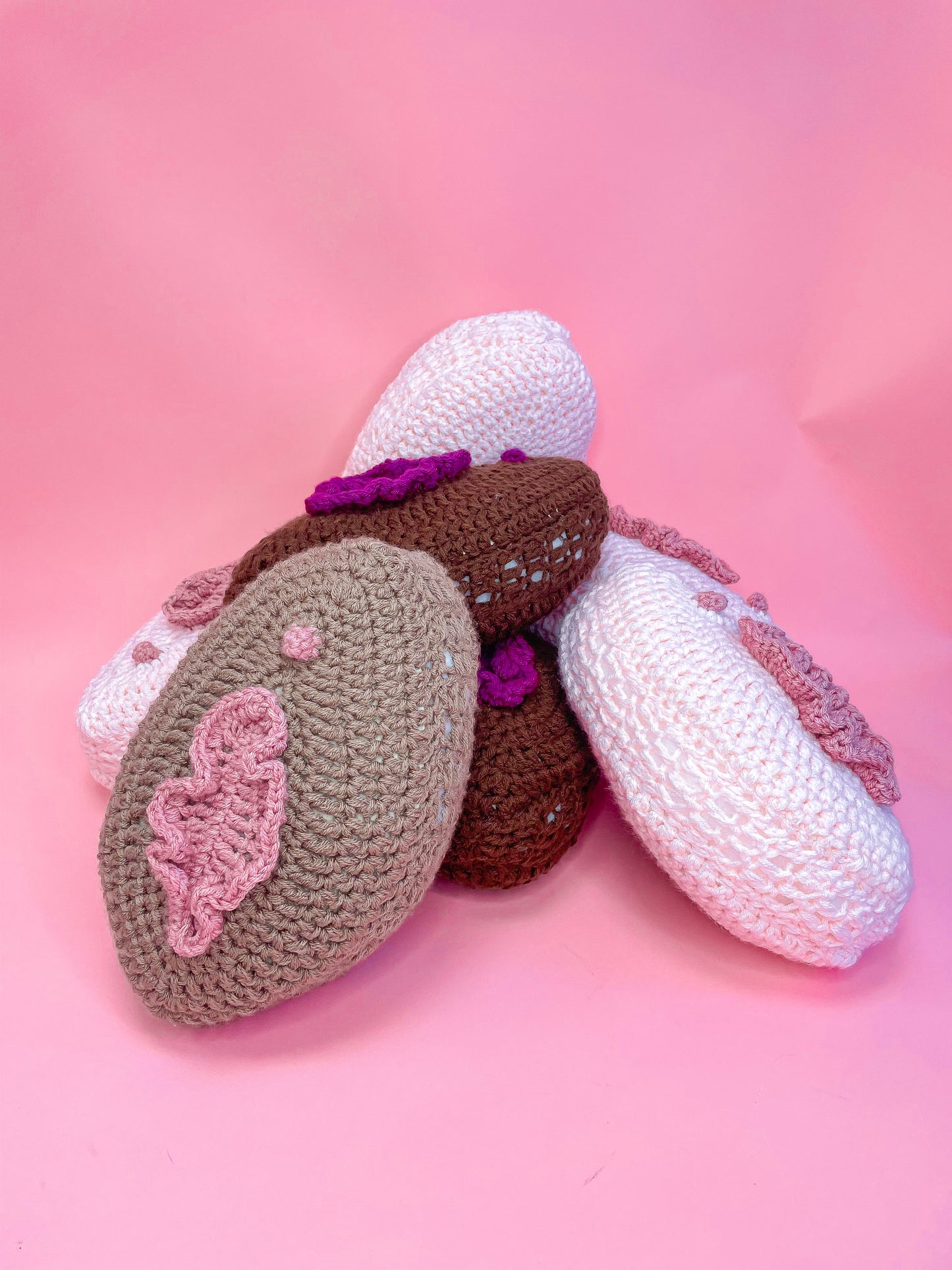 Seven crocheted cushions resembling vulvas in various colours lying on top of each other in a pile on a pink background.