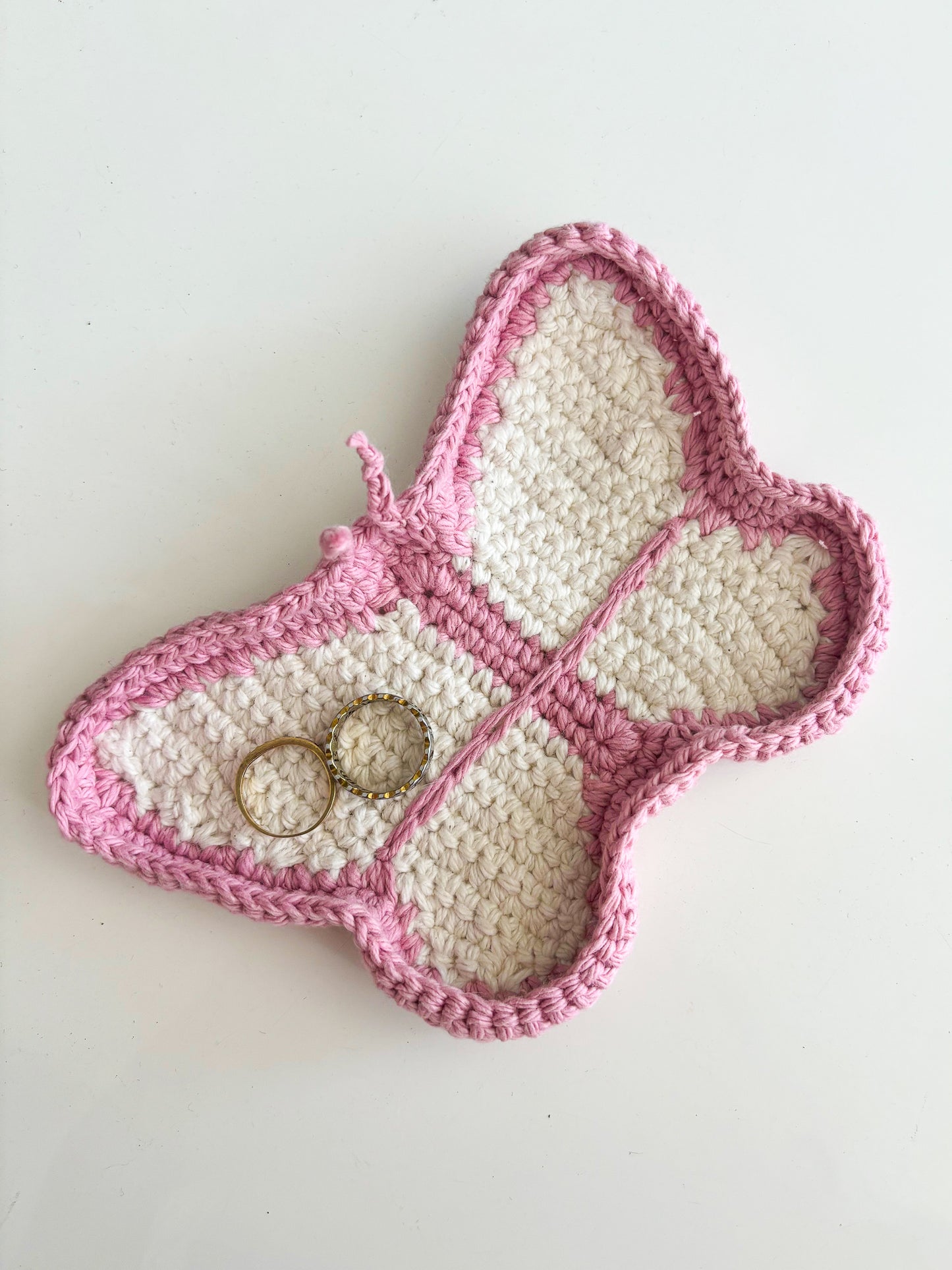 A crocheted jewellery dish shaped like a butterfly in cream and pink with some rings lying in it.