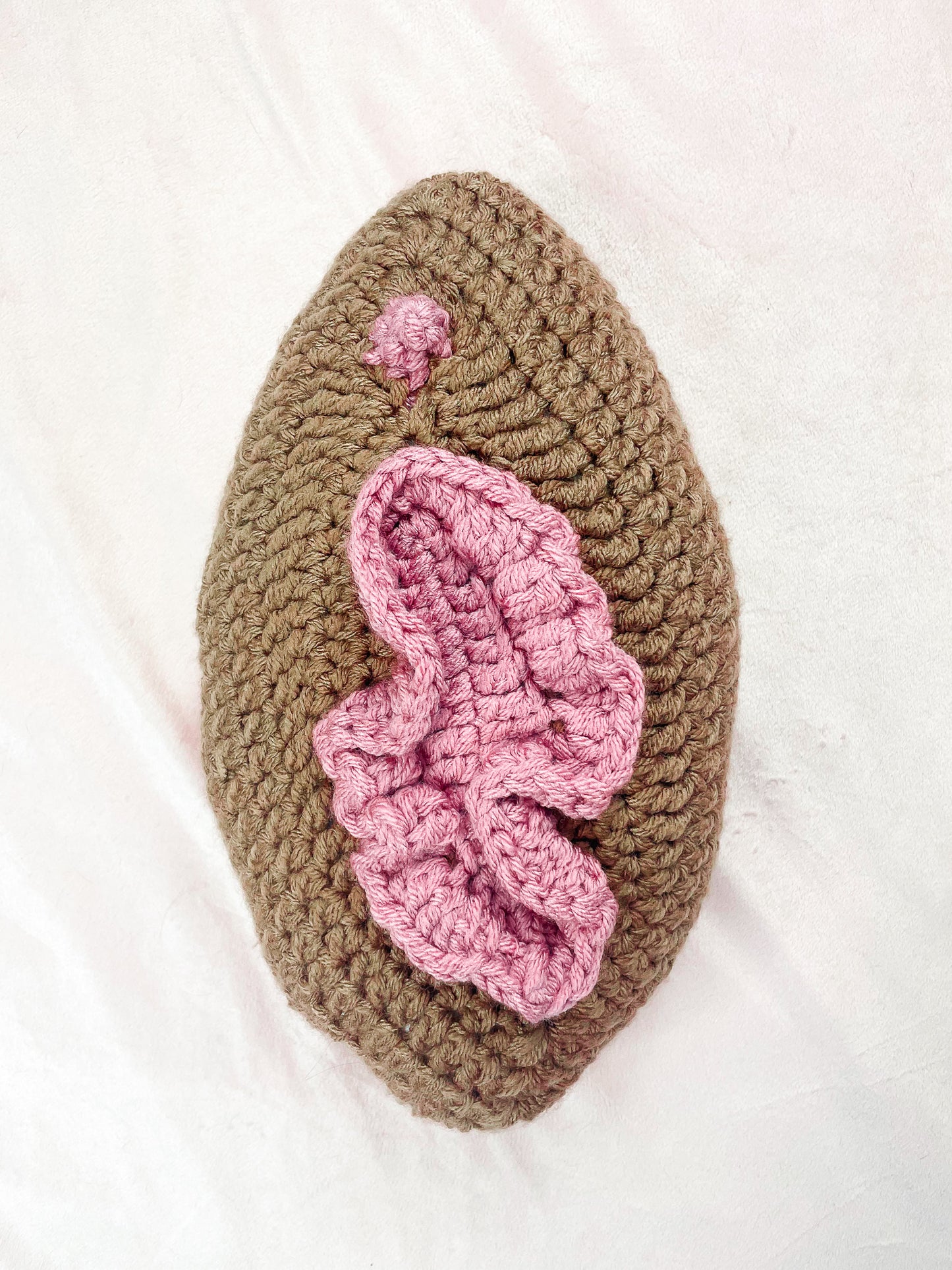 Flat lay close up of a brown cushion lying on a cream background showing the crochet stitches and 3D details resembling a vulva.