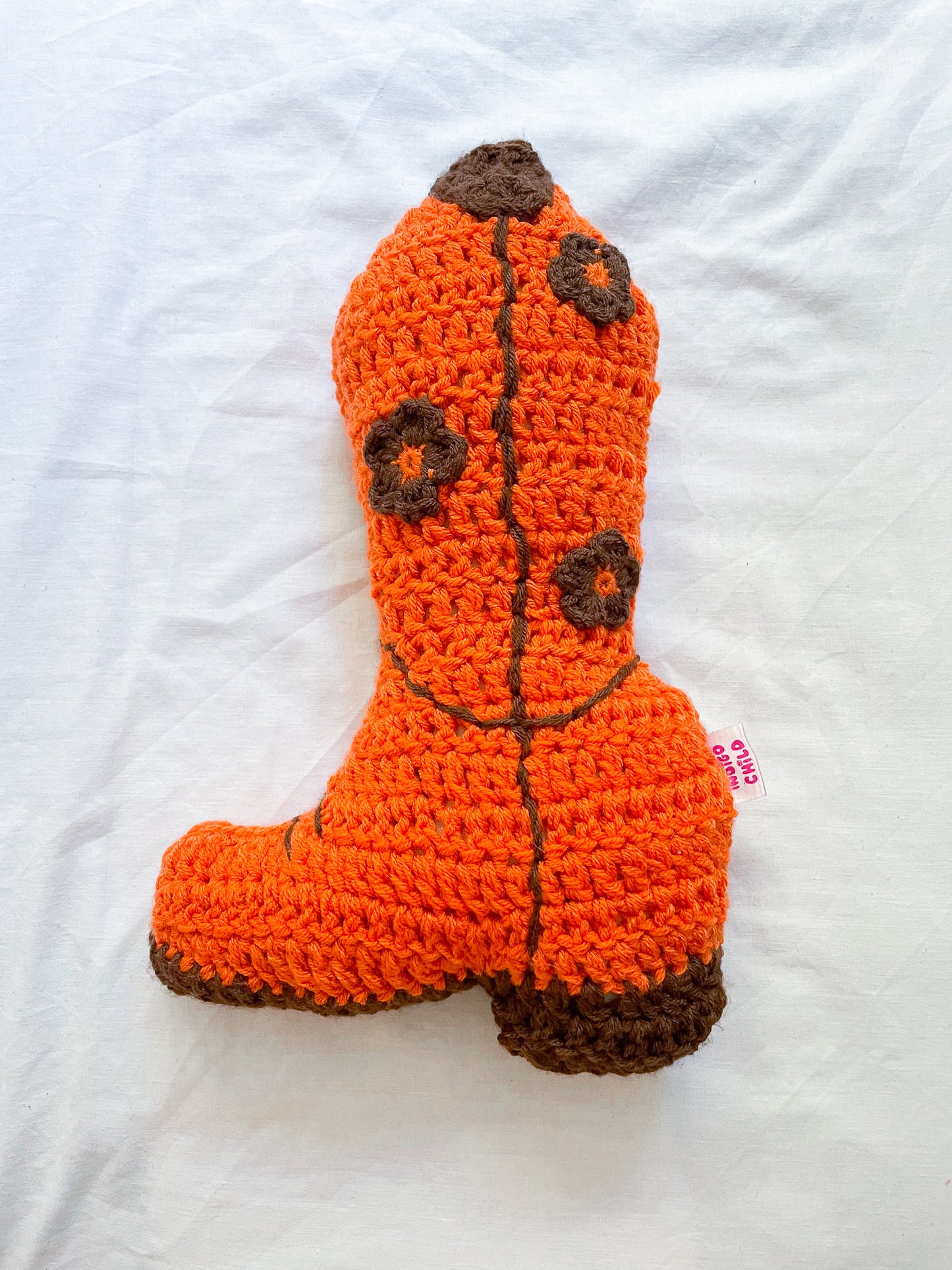 An orange crochet pillow shaped like a cowboy boot with brown detailing and brown appliqué flowers attached, lying on a white sheet.
