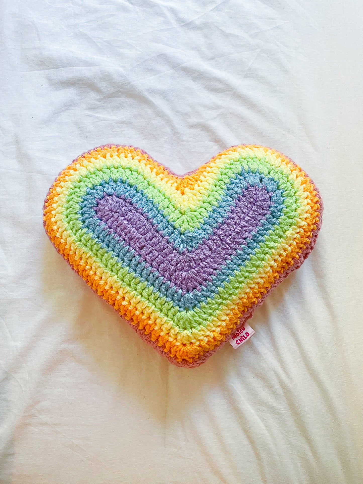 Flat lay of a pastel rainbow cushion shaped like a heart lying on a white sheet.