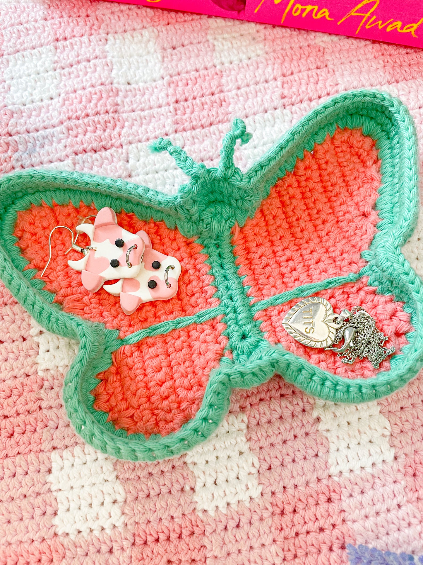 Close view of a butterfly trinket dish in pink and blue with a pair of earrings and a necklace sitting inside it lying on a pink gingham background.
