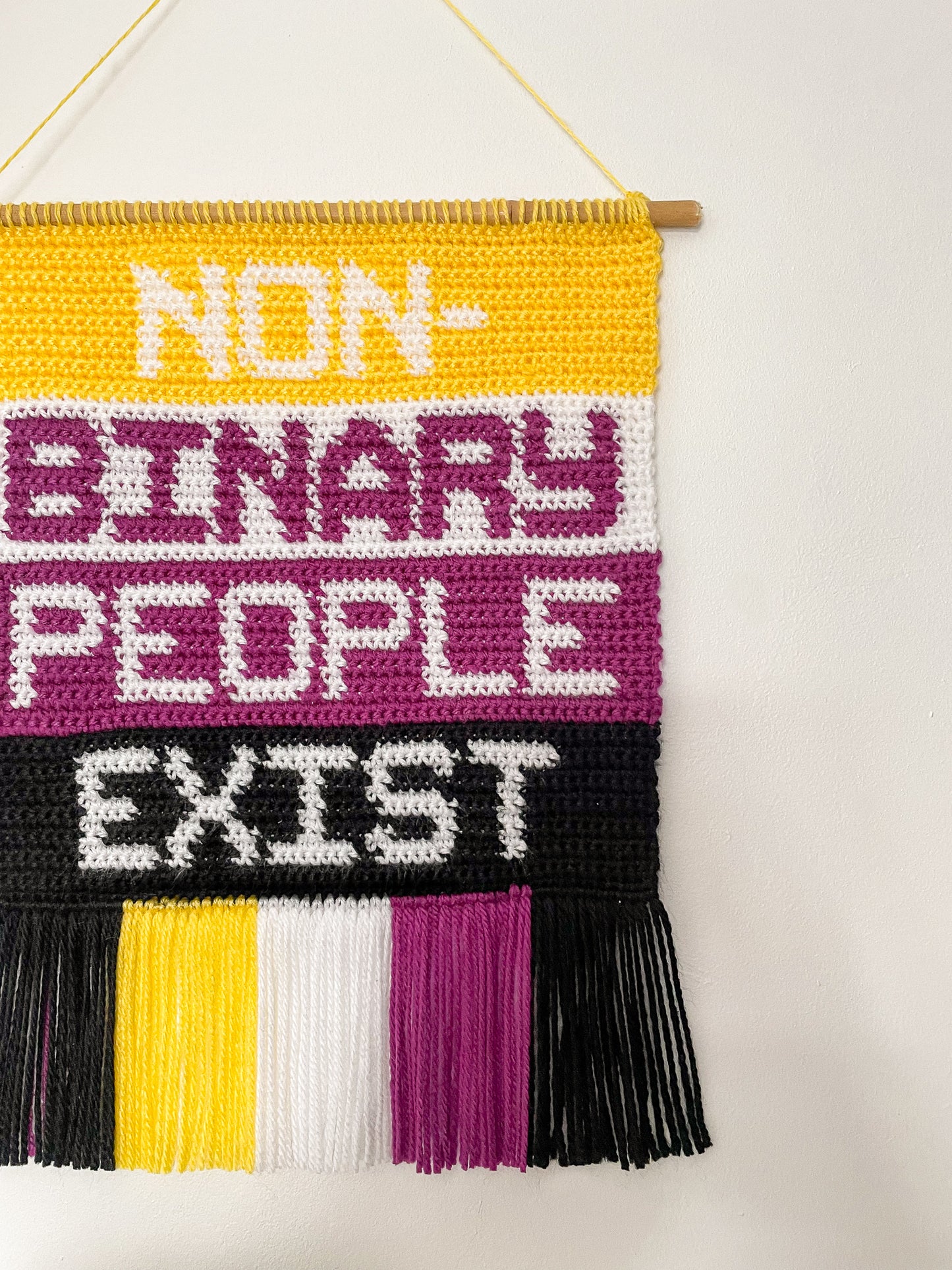Side view of a crochet wall hanging in non-binary flag colours with the words 'non binary people exist' on the front hung on a wall.
