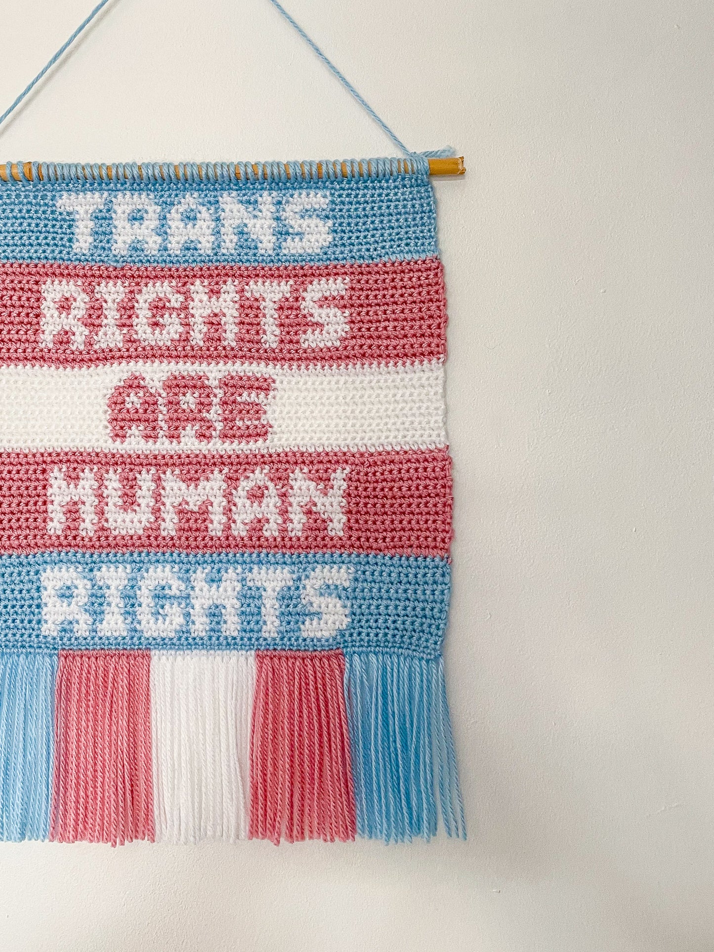 Side view of a crochet wall hanging with the quote 'trans rights are human rights' hung on a white wall.