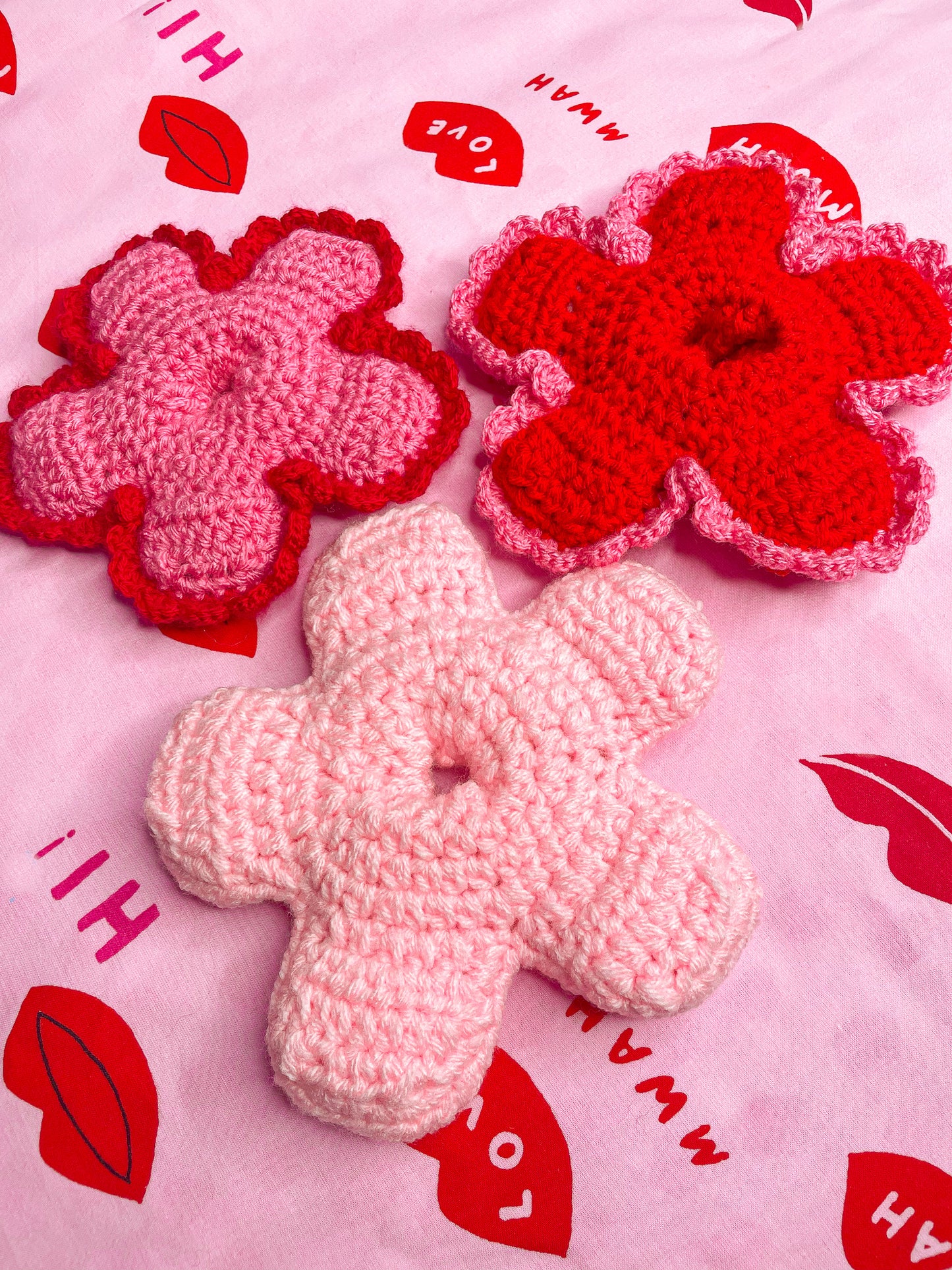 Side angle of three pink and red toned hair ties shaped like flowers lying together on a pink and red background.