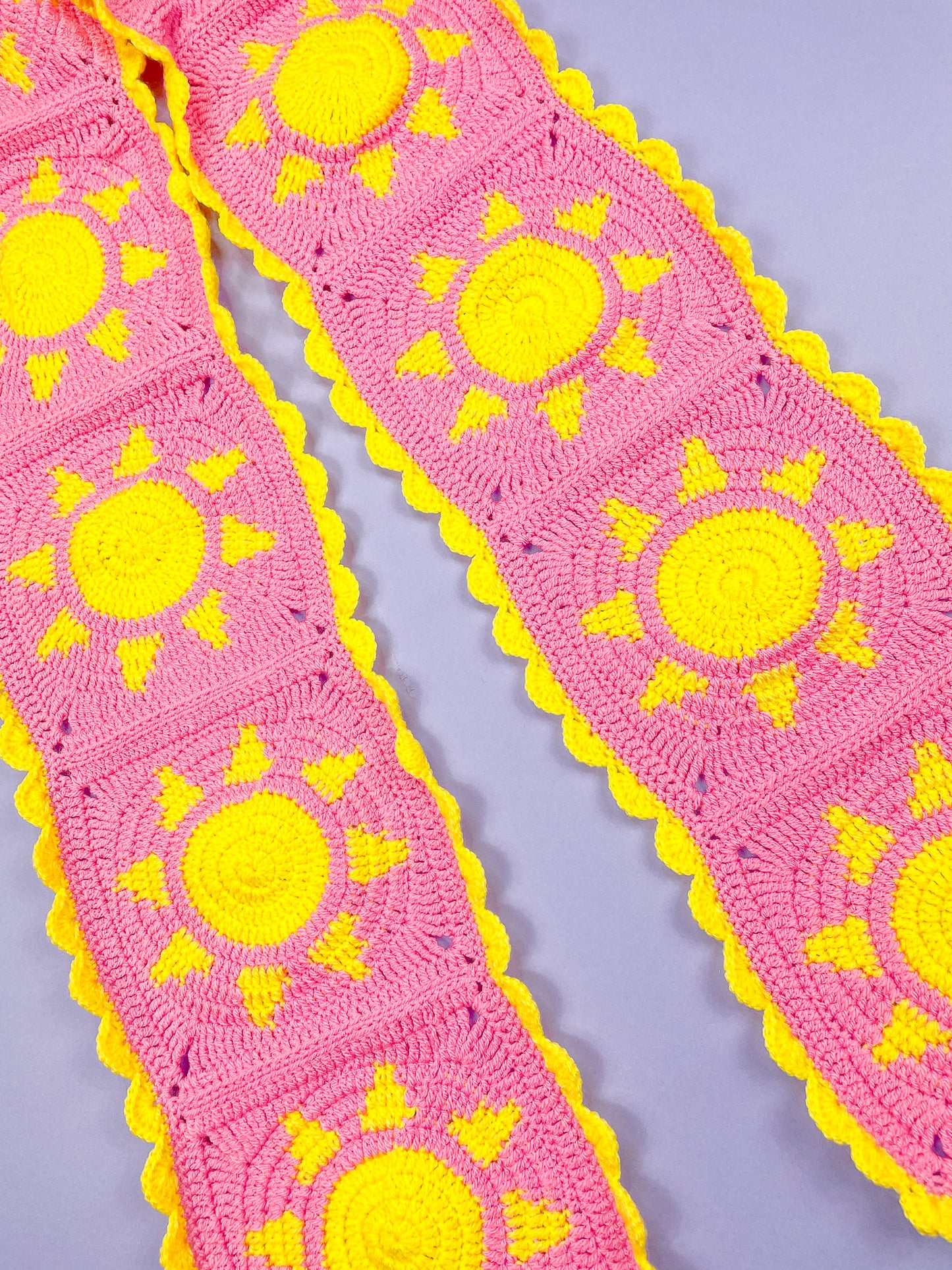 Close view of a pink scarf made up of crochet granny squares with a yellow sun design and frilled edge.