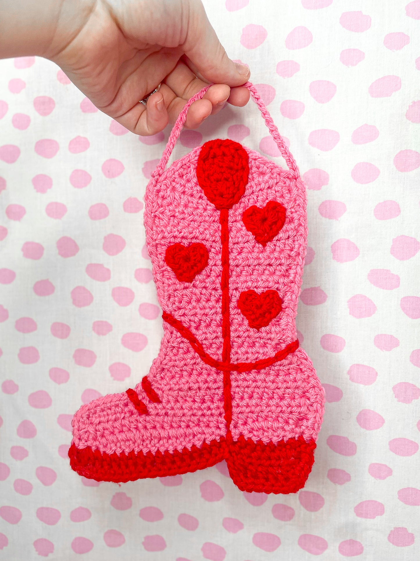 A hand holding a pink and red crochet tapestry shaped like a cowboy boot infant of a white and pink dotted background.
