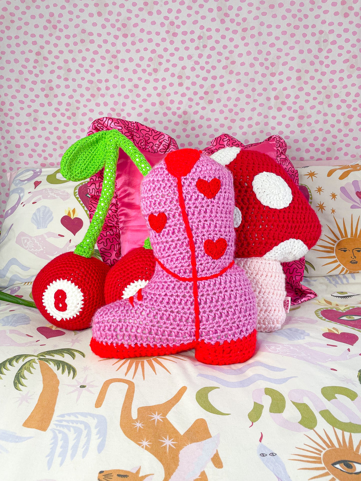 Three crocheted pillows in different shapes (cherry, cowboy boot and mushroom) lying up right together on a bed with multicoloured patterned sheets.