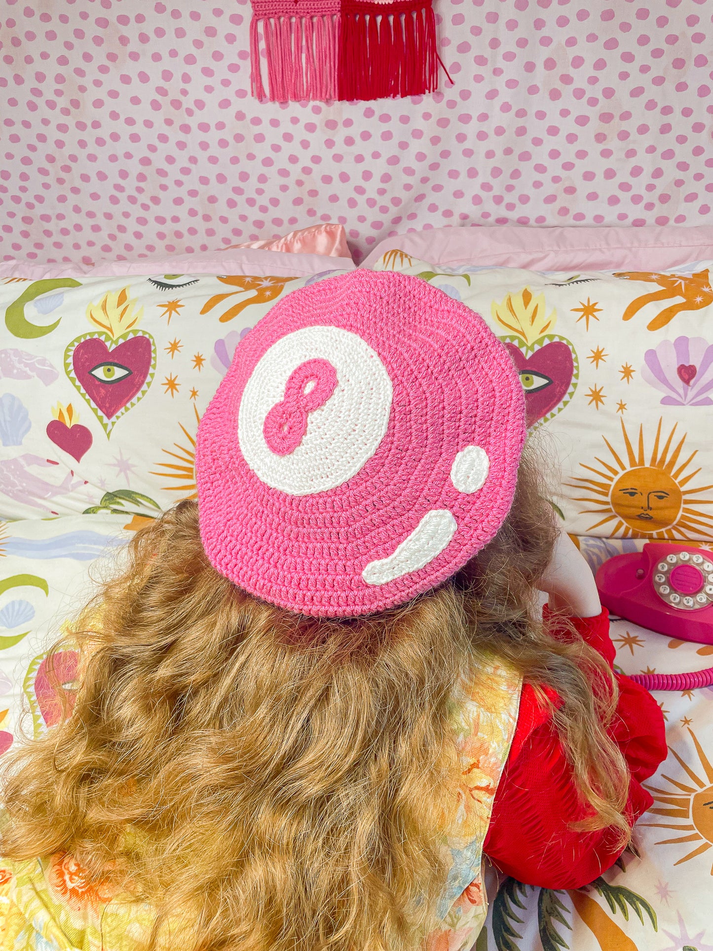 Back view of a woman lying on a bed on her front while wearing an 8 ball beret hat in pink on her head and holding a vintage phone to her ear.