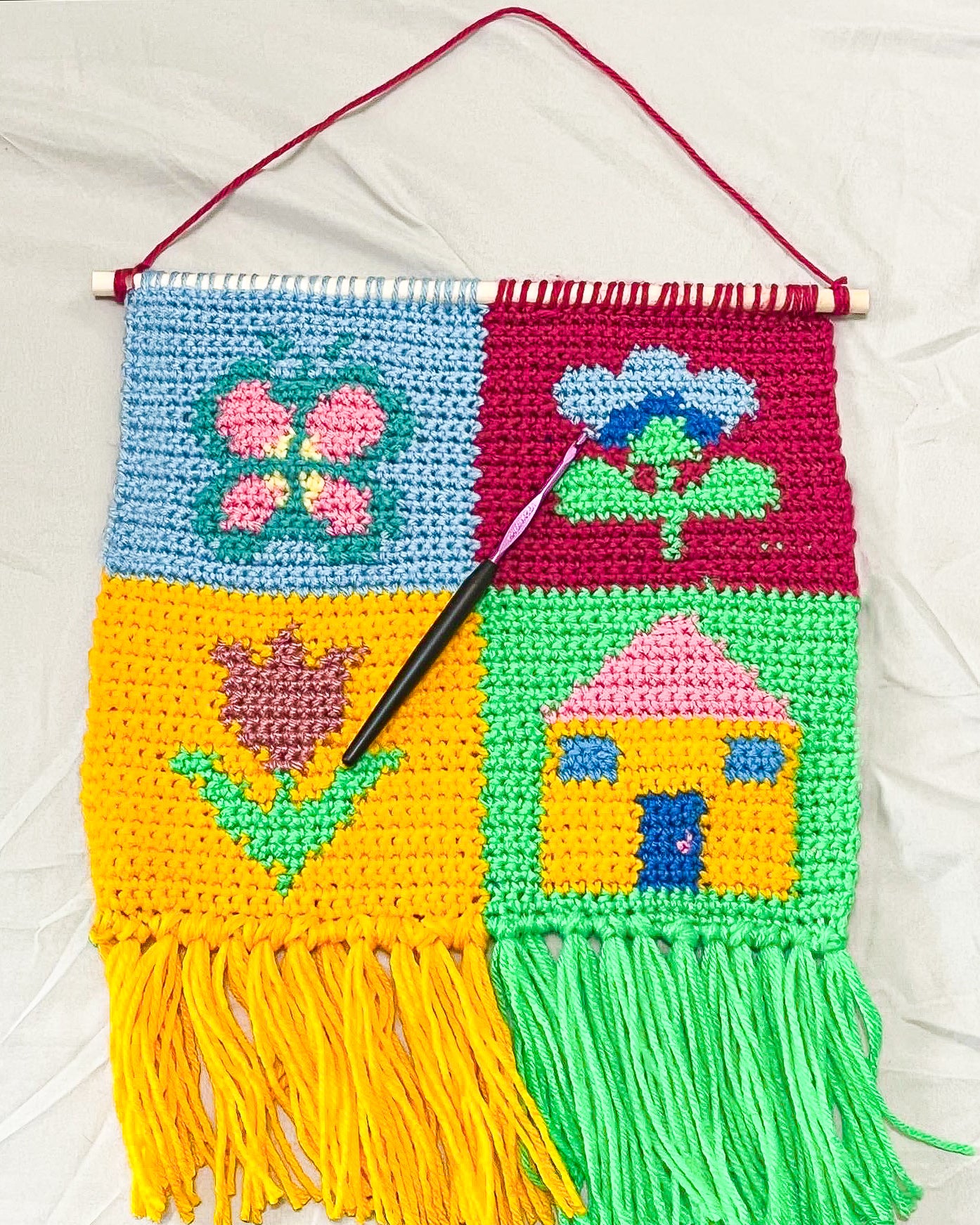 Flat lay of a pat work wall hanging in blue, burgundy, green and pink with a design of a butterfly, house and two flowers lying on a white sheet.