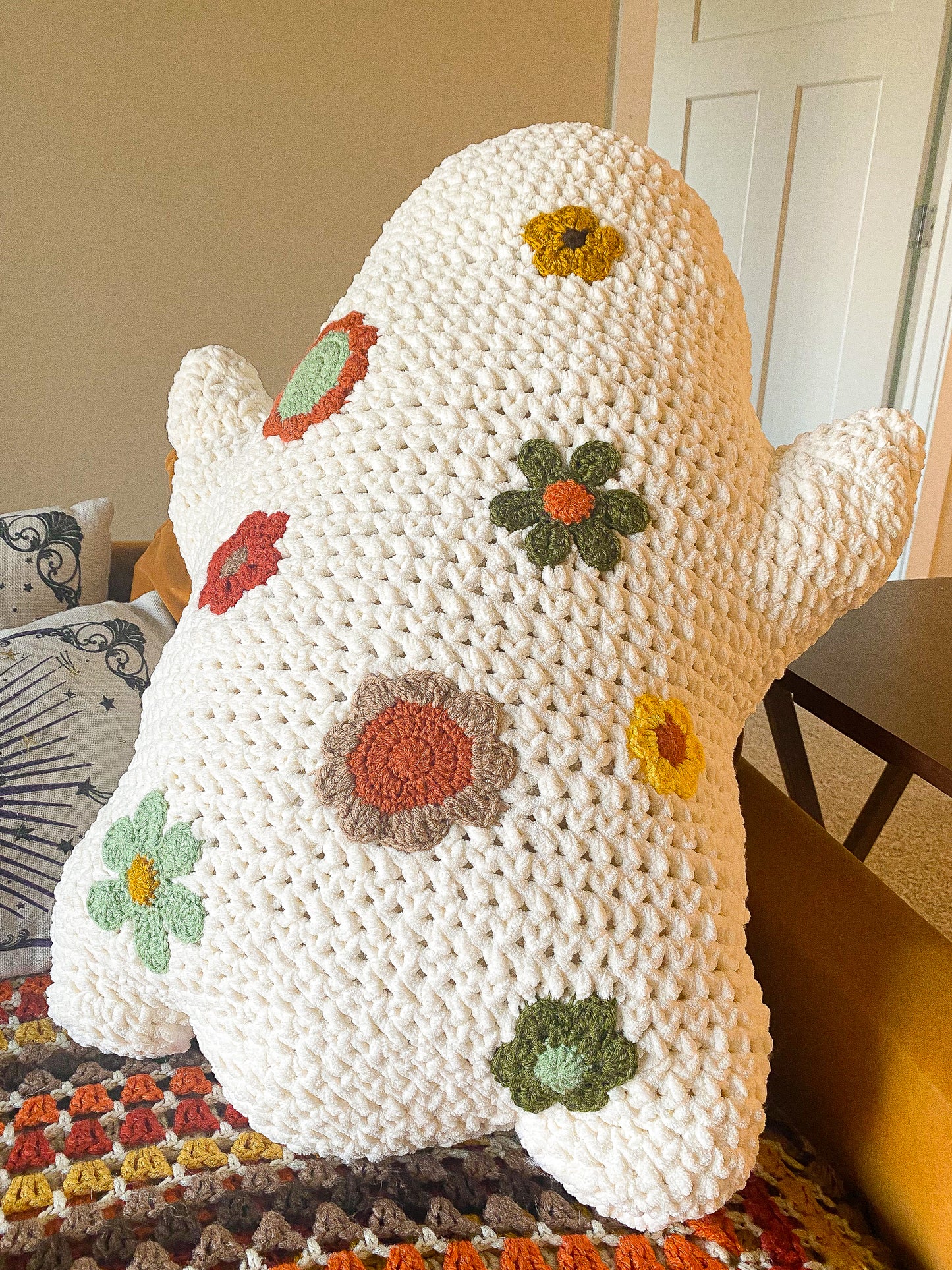 Back view of a crochet ghost shaped pillow with neutral toned flowers attached to it sitting on a tanned sofa with a matching blanket beneath it.