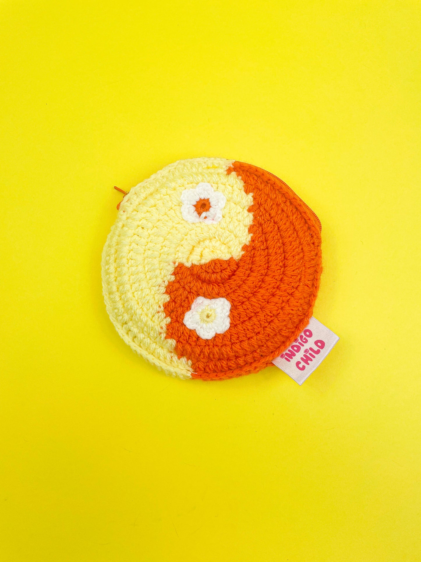 Flat lay of a circle coin purse in orange and yellow resembling a yin yang with crochet appliqué flowers for the yin yang dots, lying on a yellow background.
