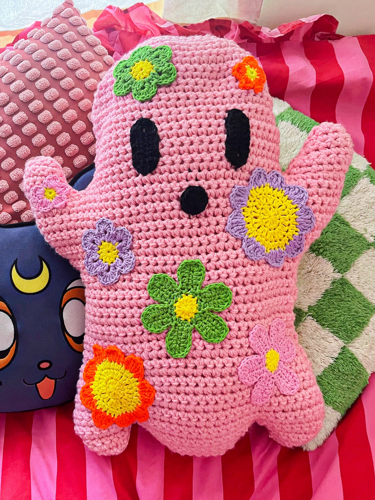 A ghost pillow in pink with bright coloured flowers sewn on, sitting with various bright coloured cushions and a pink and red striped duvet.