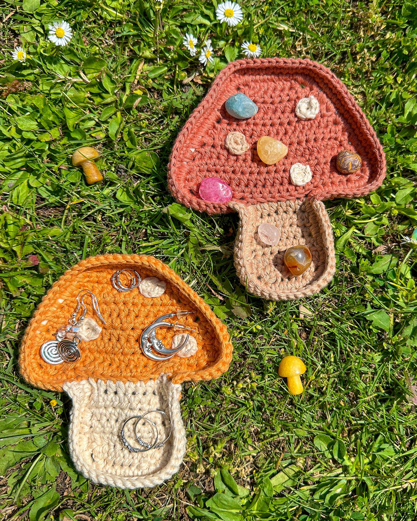 Two crocheted ring dishes in neutral colours with earrings and crystals inside them lying together on the grass. 