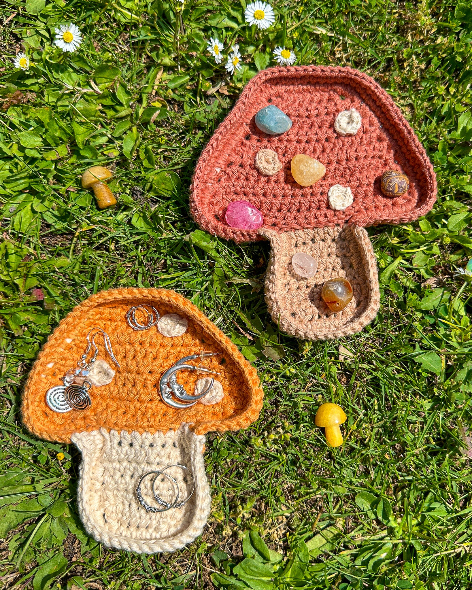 Two crocheted ring dishes in neutral colours with earrings and crystals inside them lying together on the grass. 