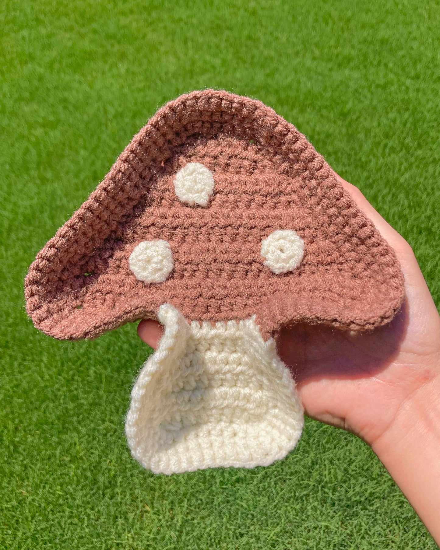 A hand holding a crocheted trinket dish shaped like a mushroom in brown and cream with grass in the background.