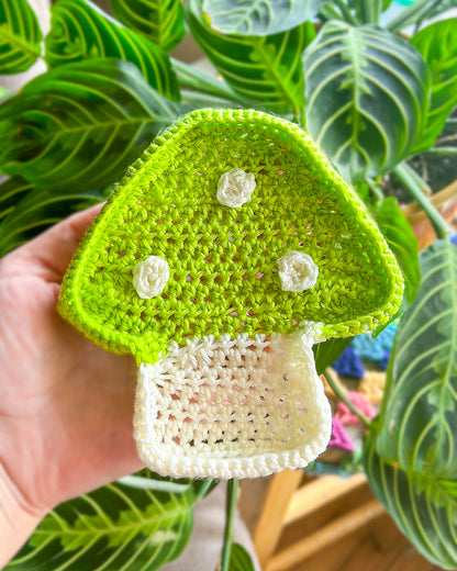 A hand holding a mushroom shaped ring dish in green and cream in front of a large plant.