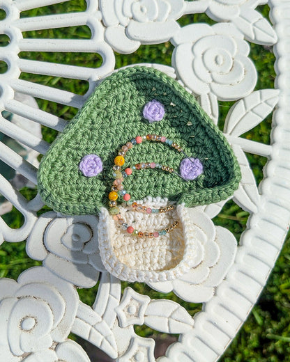 A trinket dish shaped like a mushroom in green with lilac dots with earrings in it lying on a white metal table.