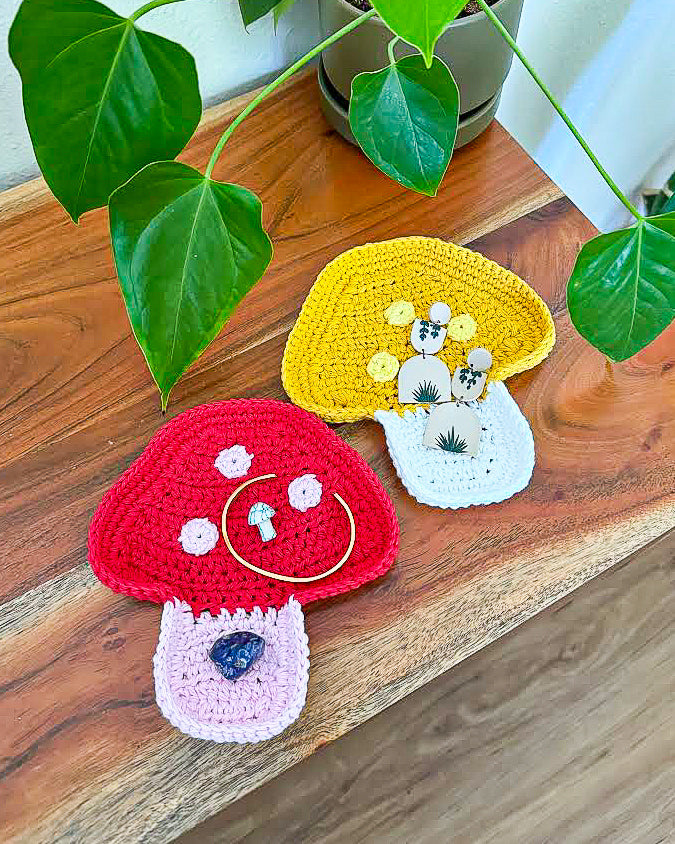 Two crocheted jewellery trays shaped like mushrooms, one red and one yellow sitting on a wooden side table with a plant next to them.