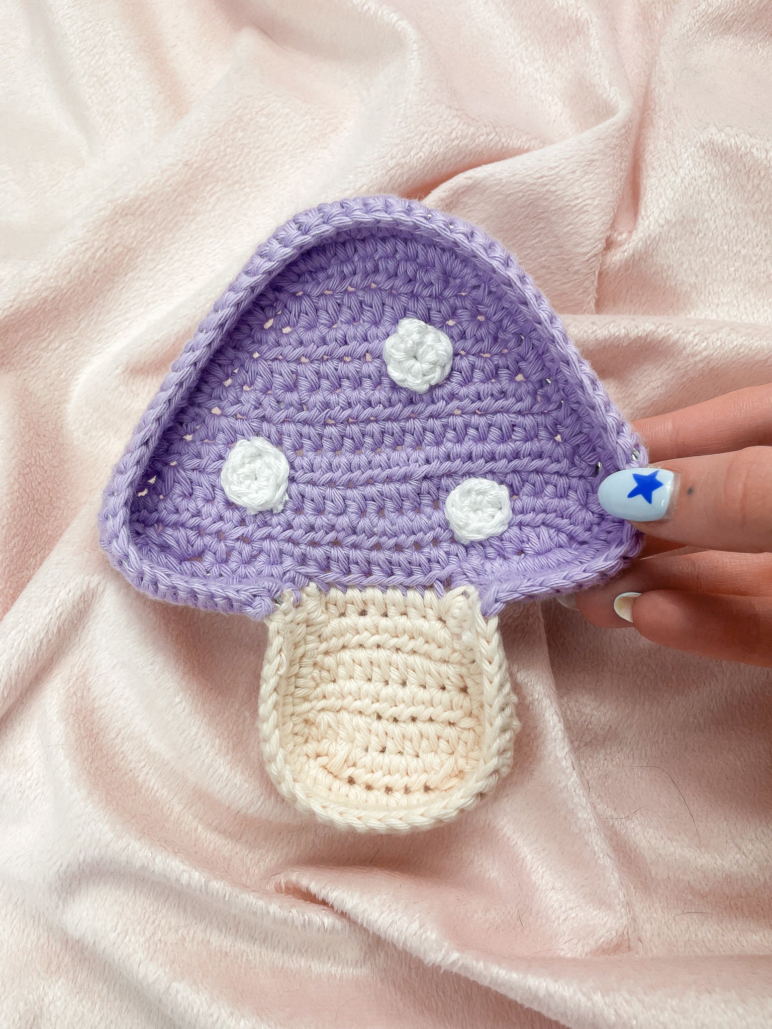 A hand holding a mushroom shaped trinket dish in lilac in front of a pale pink velvet background.
